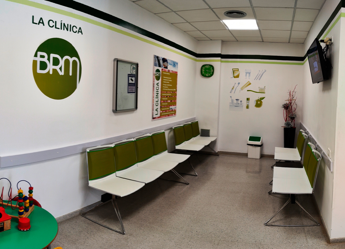 Sala d’espera Sala d'espera on hi ha cadires blanques amb un detall verd, una papelera, una taula verda on al sobre hi ha jocs per a nens, una televisió penjada a la paret esquerra, un rellotge i un cartell gran amb diferents objectes que utilitzen els dentistes, i a la paret dreta, un cartell amb el logotip de La Clínica BRM i un cartell amb els serveis que ofereix la clínica.