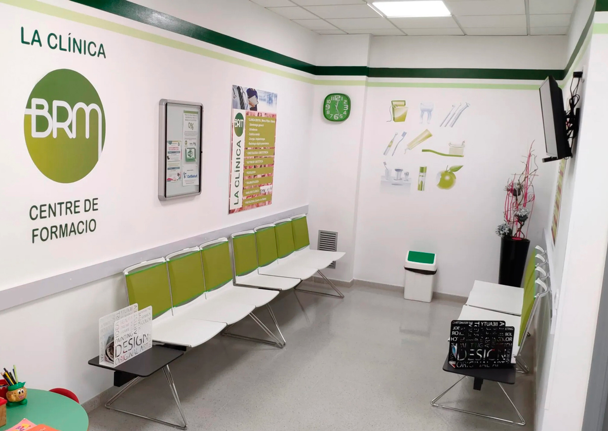 Sala d'espera on hi ha cadires blanques amb un detall verd, una papelera, una taula verda on al sobre hi ha jocs per a nens, una televisió penjada a la paret esquerra, un rellotge i un cartell gran amb diferents objectes que utilitzen els dentistes, i a la paret dreta, un cartell amb el logotip de La Clínica BRM i un cartell amb els serveis que ofereix la clínica.