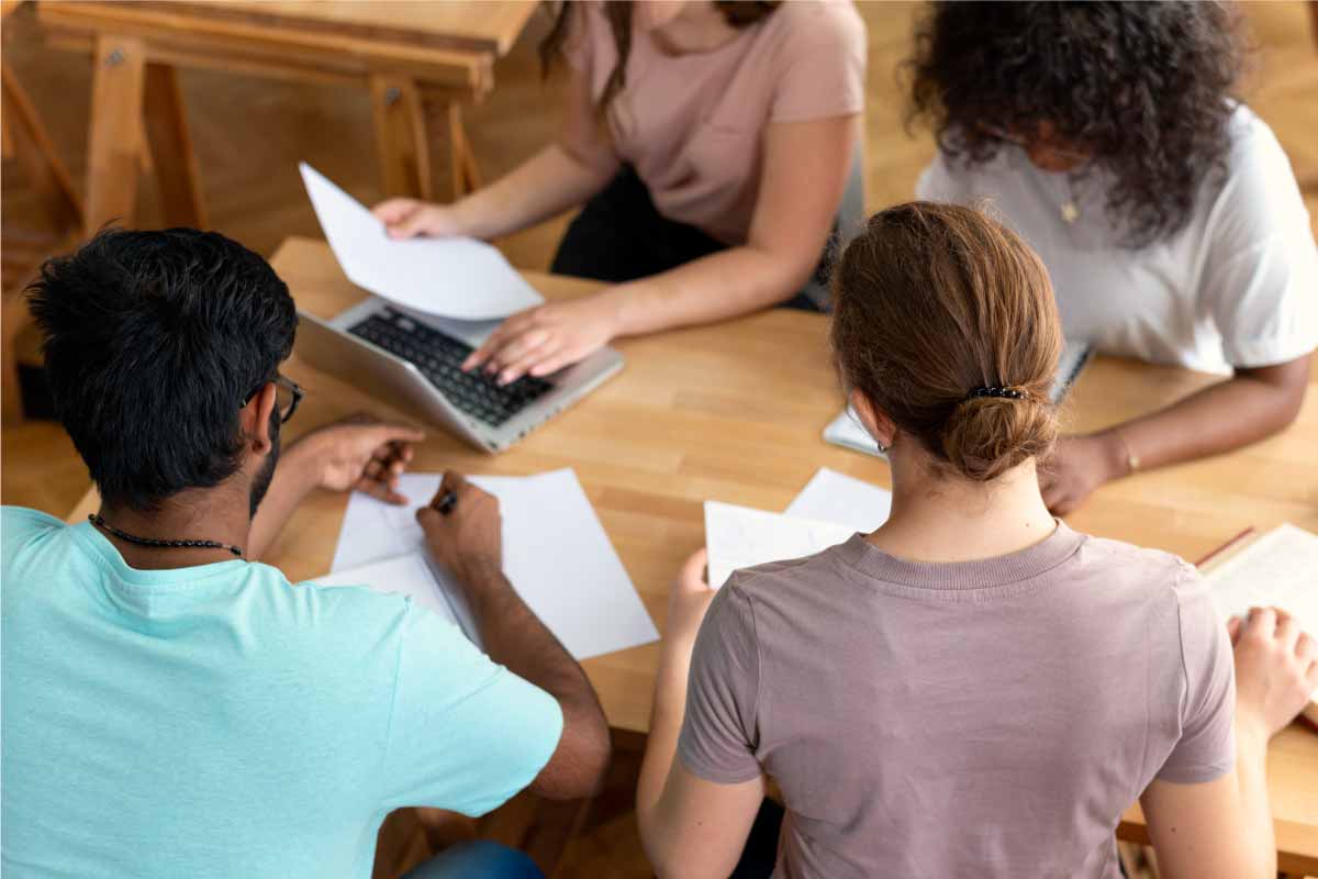 Quatre alumnes asseguts amb papers sobre la taula i un ordinador portàtil.